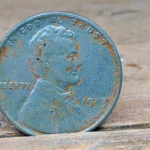 Puede incluir: Primer plano de un centavo Lincoln de 1943. La moneda es de color gris azulado con zonas de óxido. Las palabras "IN GOD WE TRUST" y "LIBERTY" son visibles, junto con el año 1943.