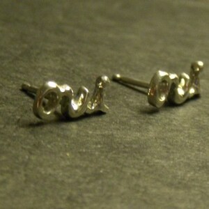 May include: A pair of silver-toned stud earrings spelling out the word "love" in a cursive script. The earrings are small and delicate, with a polished finish, and are set against a dark, textured background.