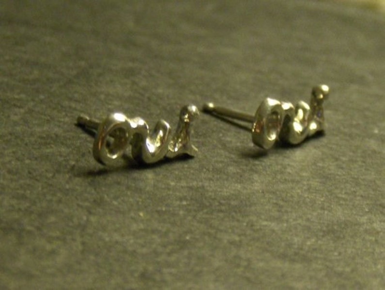 May include: A pair of silver-toned stud earrings spelling out the word "love" in a cursive script. The earrings are small and delicate, with a polished finish, and are set against a dark, textured background.