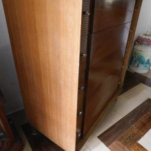 Peut inclure: Commode en bois de style moderne du milieu du si&egrave;cle. Elle pr&eacute;sente un panneau lat&eacute;ral en bois clair &agrave; grain vertical et une section de tiroirs brun fonc&eacute;. La commode comporte quatre tiroirs avec des boutons ronds et transparents et repose sur de courts pieds effil&eacute;s.