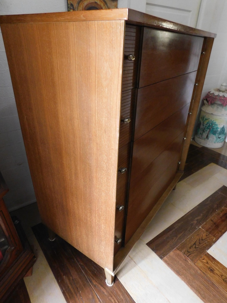 Peut inclure: Commode en bois de style moderne du milieu du si&egrave;cle. Elle pr&eacute;sente un panneau lat&eacute;ral en bois clair &agrave; grain vertical et une section de tiroirs brun fonc&eacute;. La commode comporte quatre tiroirs avec des boutons ronds et transparents et repose sur de courts pieds effil&eacute;s.