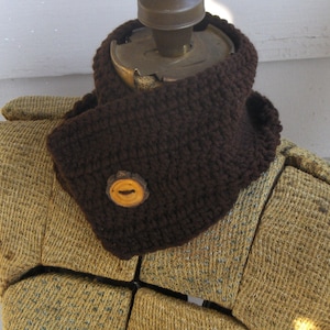 May include: A brown crocheted neck warmer with a wooden button. The neck warmer is displayed on a brown fabric covered mannequin.
