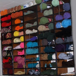May include: A wooden shelf unit filled with various colors of yarn. The yarn is organized by color and is displayed in rows on the shelves.