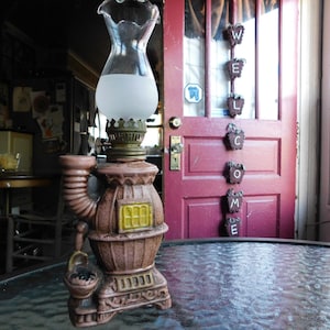 May include: A brown ceramic oil lamp shaped like a wood-burning stove with a glass chimney. The lamp is sitting on a glass table in front of a red door with a wooden sign that says "WELCOME".