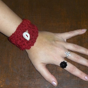 May include: A red crocheted bracelet with a white button and a white shell accent. The bracelet is worn on a person's wrist. The person is also wearing two rings on their fingers, one with a black design and the other with a silver ankh symbol.