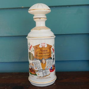 May include: White ceramic bottle with a gold lid and a colorful illustration of the North Carolina Bicentennial, 1776-1976. The illustration includes a scene of a colonial town, a document, and a red cardinal.