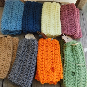 May include: Assortment of ten crocheted wrist warmers in various colors, including blue, green, orange, brown, gray, pink, and yellow. The wrist warmers are displayed on a wooden surface.