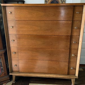 Peut inclure: Une commode en bois de style moderne du milieu du si&egrave;cle, avec six tiroirs. La commode a une finition marron clair avec un grain de bois plus fonc&eacute; sur les fa&ccedil;ades des tiroirs. Elle est dot&eacute;e de poign&eacute;es en laiton et de pieds effil&eacute;s.