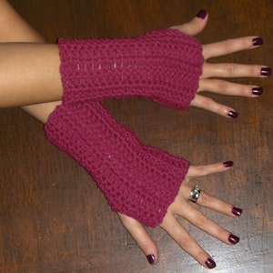 May include: A pair of burgundy fingerless gloves made of crocheted yarn. The gloves are worn on a person's hands, with the fingers exposed.