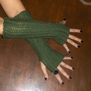 May include: A pair of green fingerless gloves made of crocheted yarn. The gloves are worn on a person's hands, and the person is wearing black nail polish.