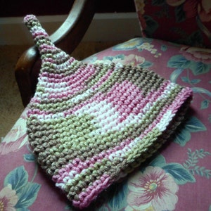 Puede incluir: Un gorro de crochet con rayas rosas, blancas y verdes. El gorro está sobre una tela con estampado floral.