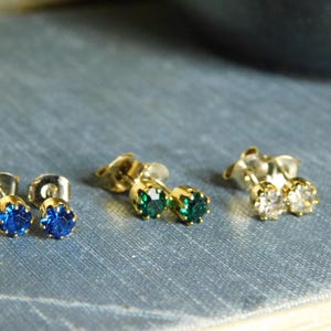 以下が含まれることがあります： さまざまな色の宝石が付いたスタッドピアスが3組。青い宝石、緑の宝石、透明な宝石のペアがあります。ピアスは金色のセッティングで、青色の背景に配置されています。