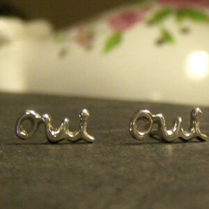 May include: A pair of silver-toned earrings spelling out the word "oui" in a cursive font. The earrings are displayed on a dark surface, with a blurred background featuring floral patterns.