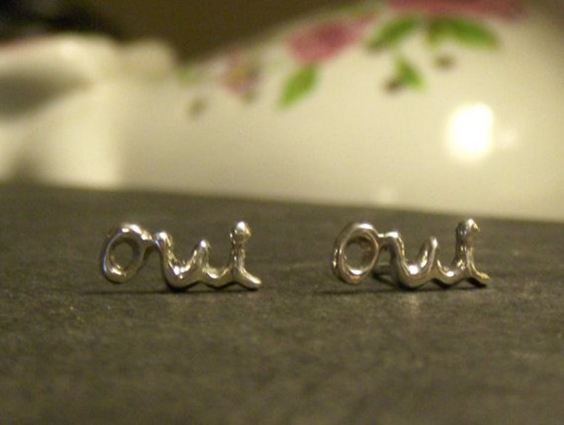 May include: A pair of silver-toned earrings spelling out the word "oui" in a cursive font. The earrings are displayed on a dark surface, with a blurred background featuring floral patterns.