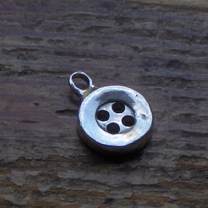 May include: A silver charm in the shape of a button with five holes. The charm has a loop at the top for attaching to a bracelet or necklace.