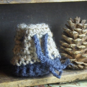May include: A small, handmade crochet bag in shades of beige, brown, and navy blue. The bag has a drawstring closure and is sitting on a wooden surface. A large pine cone is next to the bag.