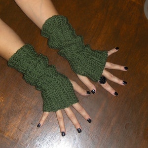 May include: A pair of green fingerless gloves with a textured knit pattern. The gloves are worn on a person's hands, with black nail polish on the fingers.