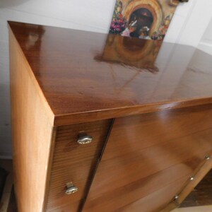 Peut inclure: Une commode en bois vintage avec une finition marron fonc&eacute; brillante. La commode pr&eacute;sente un design angulaire unique avec des tiroirs et des boutons argent&eacute;s. La surface sup&eacute;rieure refl&egrave;te l'environnement. Le grain du bois est visible.