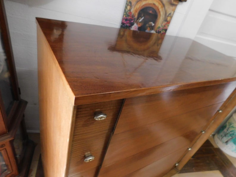 Peut inclure: Une commode en bois vintage avec une finition marron fonc&eacute; brillante. La commode pr&eacute;sente un design angulaire unique avec des tiroirs et des boutons argent&eacute;s. La surface sup&eacute;rieure refl&egrave;te l'environnement. Le grain du bois est visible.
