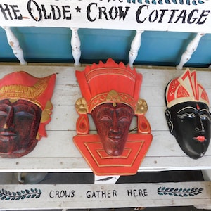 Puede incluir: Tres máscaras de madera con detalles intrincados. Las máscaras están pintadas en rojo, dorado y negro. Las máscaras se exhiben en un banco de madera blanco con las palabras "Crows Gather Here" pintadas en la parte delantera.