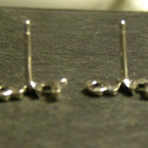 May include: Two silver-colored stud earrings with a post and a small, round backing. The earrings are set against a dark gray surface, with the posts standing upright. The earrings are likely for pierced ears.