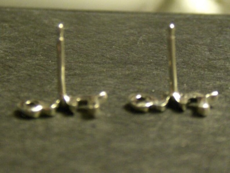 May include: Two silver-colored stud earrings with a post and a small, round backing. The earrings are set against a dark gray surface, with the posts standing upright. The earrings are likely for pierced ears.