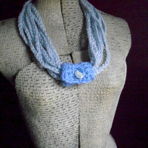 May include: A light blue crocheted necklace with a blue and white button accent. The necklace is displayed on a fabric-covered mannequin.