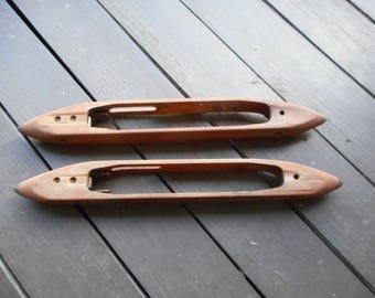 Peut inclure: Deux bobines en bois pour le tissage, avec une finition lisse et brune et une extrémité légèrement pointue. Les bobines sont posées sur une surface en bois.