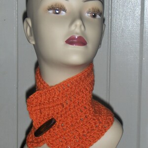 May include: A close-up of a mannequin wearing a bright orange crocheted neck warmer with a brown button. The neck warmer is folded over and has a textured pattern.