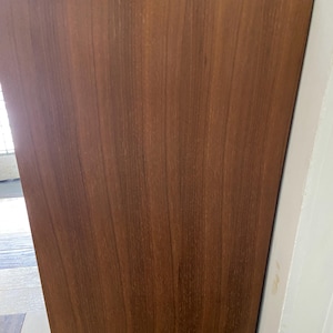 Peut inclure: Un meuble en bois de style milieu du si&egrave;cle, de couleur marron chaud. Le meuble a un design simple et rectangulaire avec un grain de bois visible. Le bord sup&eacute;rieur est l&eacute;g&egrave;rement plus fonc&eacute;, ajoutant de la profondeur. Il est plac&eacute; contre un mur blanc.