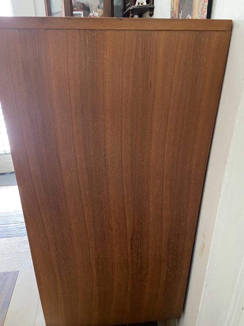 Peut inclure: Un meuble en bois de style milieu du si&egrave;cle, de couleur marron chaud. Le meuble a un design simple et rectangulaire avec un grain de bois visible. Le bord sup&eacute;rieur est l&eacute;g&egrave;rement plus fonc&eacute;, ajoutant de la profondeur. Il est plac&eacute; contre un mur blanc.