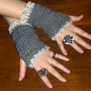 Puede incluir: Un par de guantes sin dedos de ganchillo gris y blanco con un borde de ganchillo blanco. Los guantes se llevan en las manos de una persona, y la persona lleva dos anillos en sus dedos. Un anillo es un anillo de plata con una piedra grande de color gris oscuro. El otro anillo es una banda de plata.