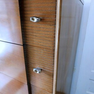 Peut inclure: Une commode en bois vintage avec une finition brun clair. Elle comporte trois tiroirs avec des d&eacute;tails de lattes horizontales et des poign&eacute;es ovales argent&eacute;es. Le c&ocirc;t&eacute; de la commode a une surface lisse et polie. Elle est plac&eacute;e contre un mur blanc.