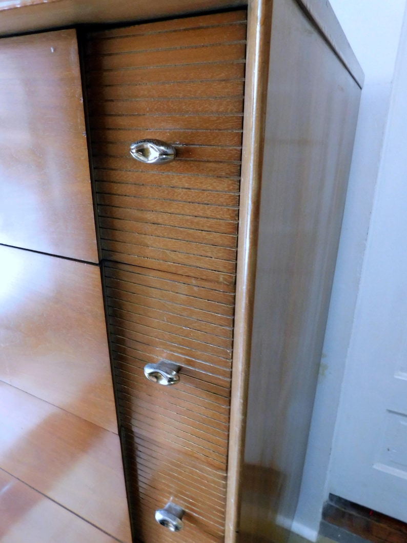 Peut inclure: Une commode en bois vintage avec une finition brun clair. Elle comporte trois tiroirs avec des d&eacute;tails de lattes horizontales et des poign&eacute;es ovales argent&eacute;es. Le c&ocirc;t&eacute; de la commode a une surface lisse et polie. Elle est plac&eacute;e contre un mur blanc.