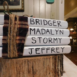Puede incluir: Pila de cuatro bloques de madera pintados de blanco con los nombres "Bridger", "Madalyn", "Stormy" y "Jeffrey" escritos con marcador negro. Los bloques están atados con cuerda y una cinta de cuadros rojos y negros.