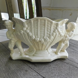 Puede incluir: Un macetero de cerámica blanca con forma de concha marina con dos querubines a cada lado. El macetero está decorado con detalles florales en relieve.