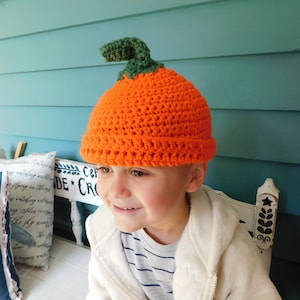 May include: A young child wearing an orange and green crocheted pumpkin hat. The hat has a stem and is made with a textured stitch.