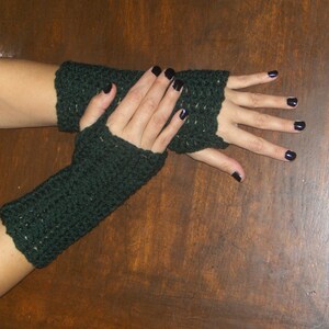 Pode incluir: Um par de luvas sem dedos de cor verde escura feitas de fio de crochê. As luvas são usadas nas mãos de uma pessoa, e as unhas da pessoa são pintadas de preto.