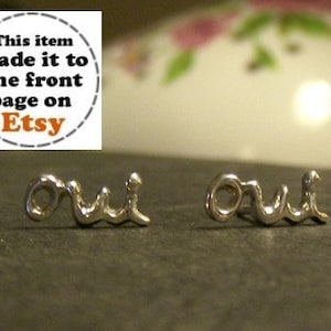 May include: A pair of silver-toned stud earrings spelling out the word "oui" in a cursive font. The earrings are displayed on a dark surface. A circular graphic in the upper left corner states "This item made it to the front page on Etsy."