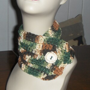 May include: A crocheted neck warmer in a camouflage pattern with brown, green, and white yarn. The neck warmer has a white button in the center.