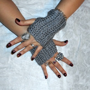 May include: A pair of gray fingerless gloves with a textured knit pattern. The gloves are worn on a person's hands, which are adorned with rings and nail polish.