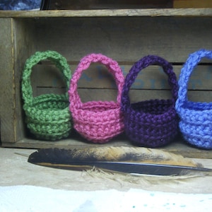 May include: Four small crocheted baskets in green, pink, purple, and blue, sitting in a wooden crate.