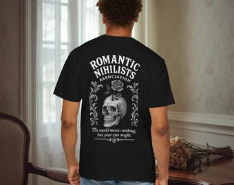 Op de afbeelding: Zwart t-shirt met een grafische print op de achterkant. Het ontwerp toont een schedel met een roos en decoratieve krullen, met de tekst "Romantic Nihilists Association". De tekst "The world means nothing, but your eyes might" staat eronder.