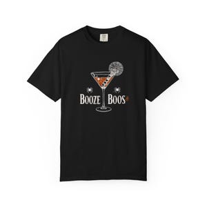 Peut inclure: T-shirt noir avec un motif sur le thème d'Halloween. Le design comprend un verre à cocktail avec une garniture en toile d'araignée, des araignées et le texte "Booze & Boos" en blanc. Un vêtement amusant et festif.
