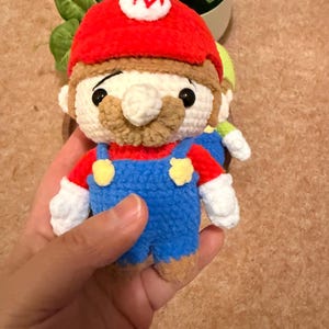 May include: Crocheted Mario doll with a red hat featuring a white "M", a tan mustache, and blue overalls. The doll has white gloves and black eyes. A second doll is partially visible in the background.