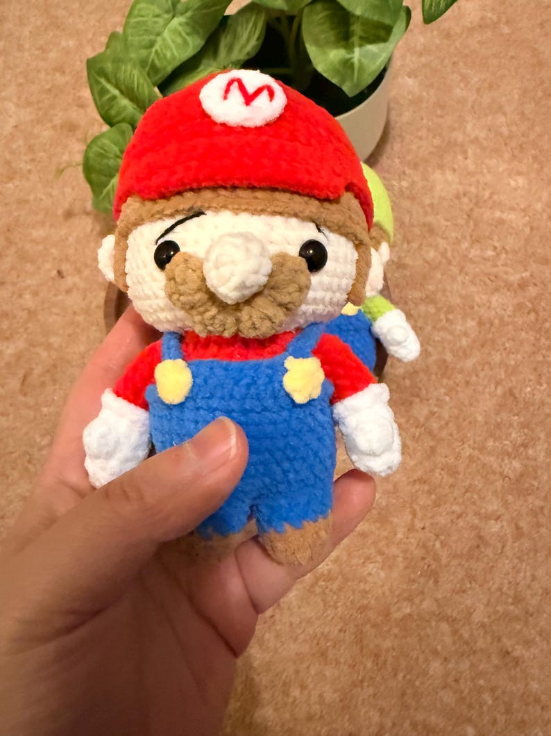 May include: Crocheted Mario doll with a red hat featuring a white "M", a tan mustache, and blue overalls. The doll has white gloves and black eyes. A second doll is partially visible in the background.
