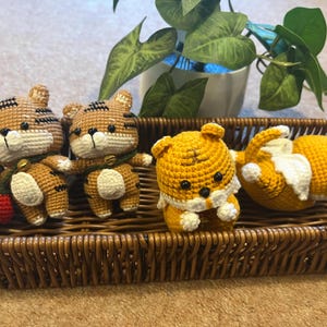 May include: A collection of four crocheted tiger figurines in a woven basket. The tigers are primarily orange and brown with white accents. Two are upright, and two are lying down. A green plant is in the background.