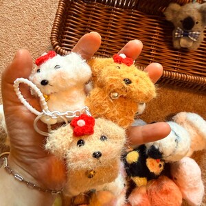 May include: A collection of handmade felt dog and koala bear keychains. The dogs are white, tan, and orange, each with a red flower and a small bell. The koala has a blue bow tie. The keychains are displayed in a brown wicker basket.