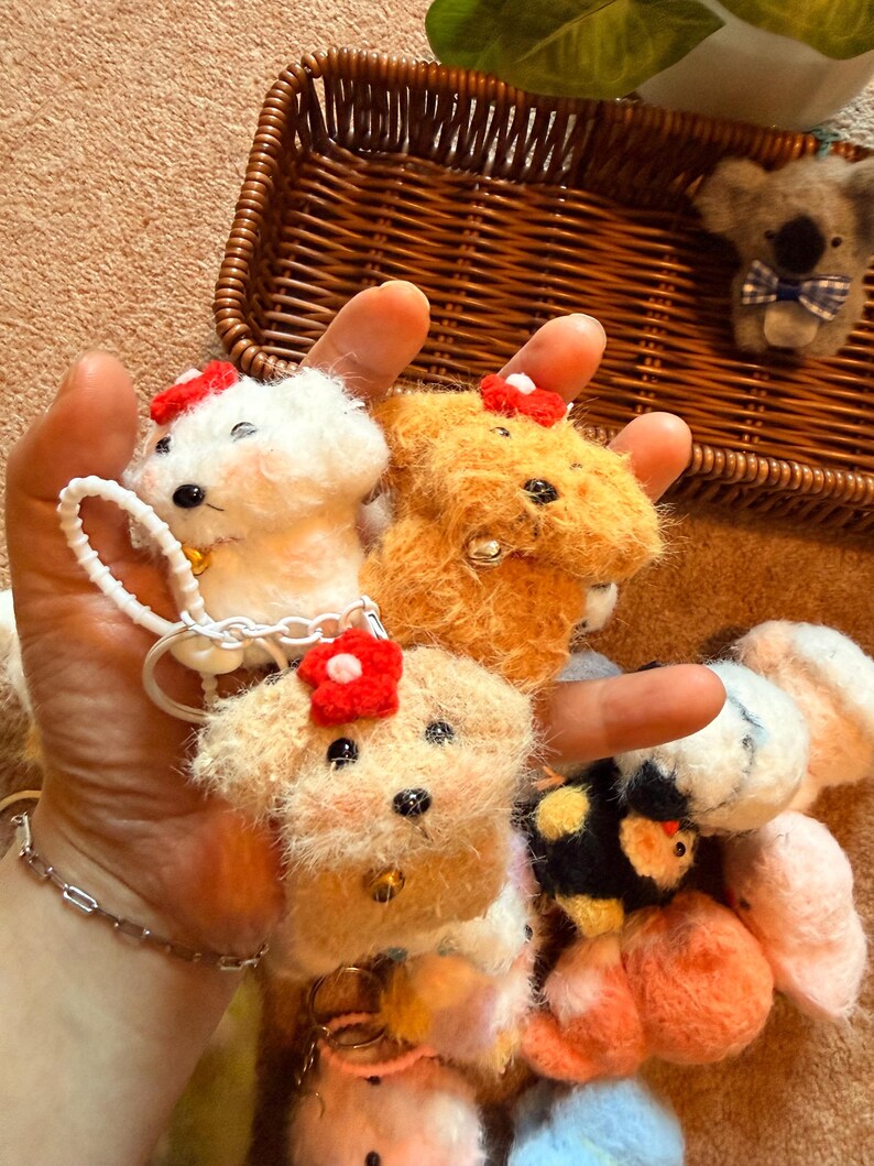May include: A collection of handmade felt dog and koala bear keychains. The dogs are white, tan, and orange, each with a red flower and a small bell. The koala has a blue bow tie. The keychains are displayed in a brown wicker basket.