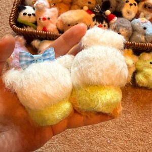 May include: A hand holding two small, fluffy, white and yellow plush toys with a blue and white checkered bow. A wicker basket filled with various small plush animals is in the background.